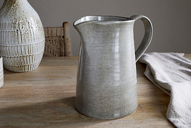 The Valinha Earthenware Jug in Grey (Large) from nkuku sits on a wooden table, bringing elegance to your serveware. Behind it, a textured vase and woven chair enhance the earthy decor, while a draped cloth softens the dining scene.