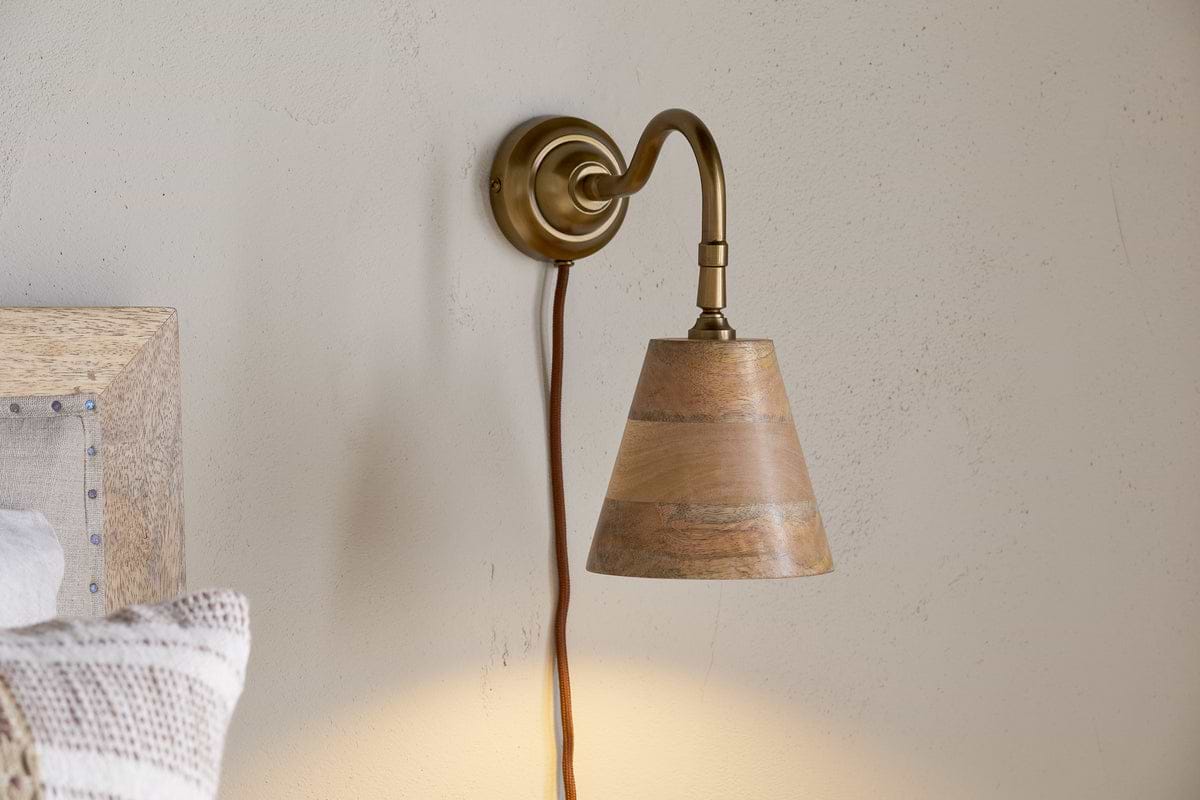 The nkuku Vattu Mango Wood Plug In Wall Lamp in Natural & Antique Brass is mounted on a beige wall, its brown cord hanging down as warm, ambient light glows. A bed with a textured pillow appears at the left edge.