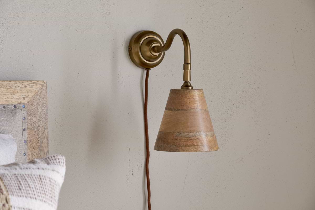 The nkuku Vattu Mango Wood Plug In Wall Lamp in Natural & Antique Brass is mounted on a beige wall, adding natural texture. A brown cord hangs beneath, and part of a wooden bed frame with a striped pillow appears at the edge.