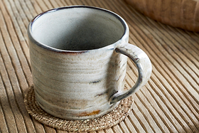 The Vizo Mug - Grey - Large by nkuku, featuring subtle brown and gray tones, rests on a round woven coaster atop a grooved wooden table.