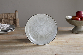 The Valinha Earthenware Dinnerware Set - Grey by nkuku features a light gray ceramic plate with circular patterns and reactive glaze, standing on a wooden table beside stacked plates, a woven chair, and a fruit bowl with red apples.