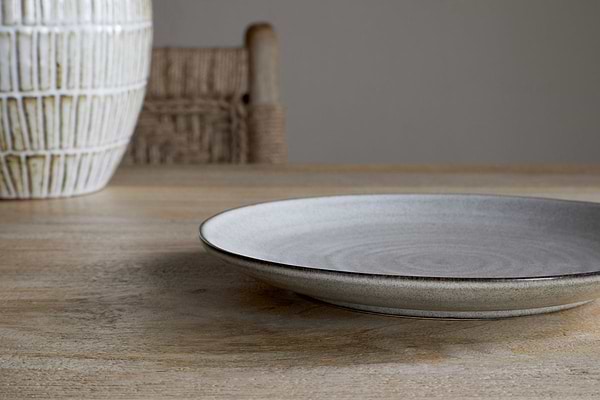 The Valinha Earthenware Side Plate - Grey by nkuku rests on a wooden table, accompanied by part of a textured vase and a woven chair in the background, creating a calm and minimalistic setting.