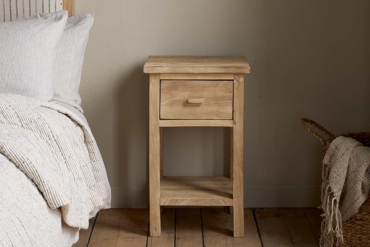 The Vivan Grooved Bedside Table - Natural by nkuku, featuring a drawer and lower shelf, stands beside a neatly made bed with light bedding on a wooden floor next to a woven basket.