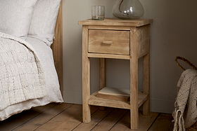 The Vivan Grooved Bedside Table - Natural by nkuku sits by a bed with white bedding. A glass and vase are on top, while two books rest neatly on the open shelf above the wooden floor.