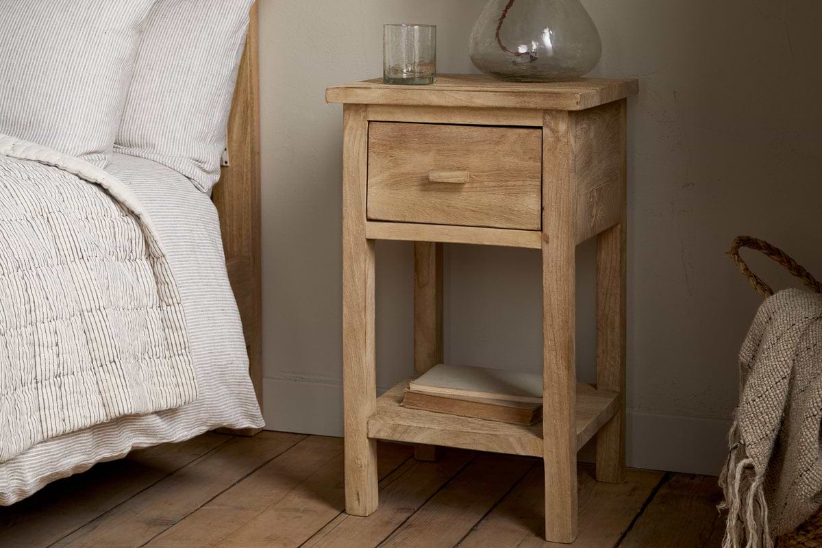 The Vivan Grooved Bedside Table - Natural by nkuku sits by a bed with white bedding. A glass and vase are on top, while two books rest neatly on the open shelf above the wooden floor.