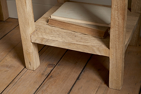 Two old books, one closed and one slightly open, rest on the lower shelf of the nkuku Vivan Grooved Bedside Table - Natural. The scene features a wooden floor with visible grain and knots.