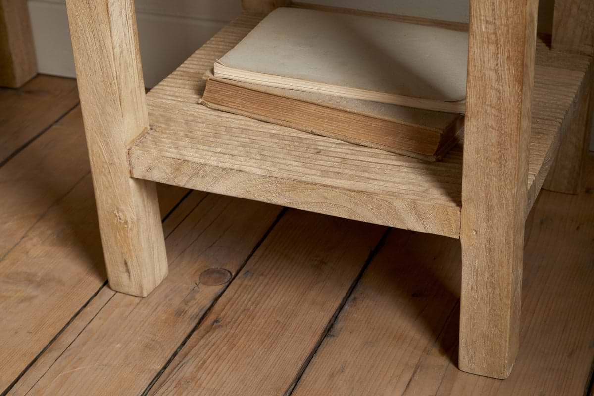 Two old books, one closed and one slightly open, rest on the lower shelf of the nkuku Vivan Grooved Bedside Table - Natural. The scene features a wooden floor with visible grain and knots.
