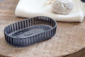 The Valeska Recycled Glass Soap Dish by nkuku, featuring a ribbed oval shape, rests on wood. A folded white towel and light pumice stone in the background add a spa-like touch to these elegant bathroom accessories.