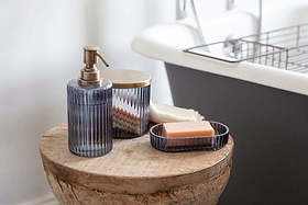 The nkuku Valeska Recycled Glass Soap Dish with an orange bar of soap, a ribbed recycled glass dispenser, and a matching lidded container rest on a rustic wooden stool beside a bath, adding cozy, modern style to the bathroom.