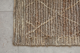 A close-up of the nkuku Vandi Jute Doormat - Natural, featuring thick, textured braids in beige and brown tones with a geometric diamond design, displayed on a light gray stone tile floor.