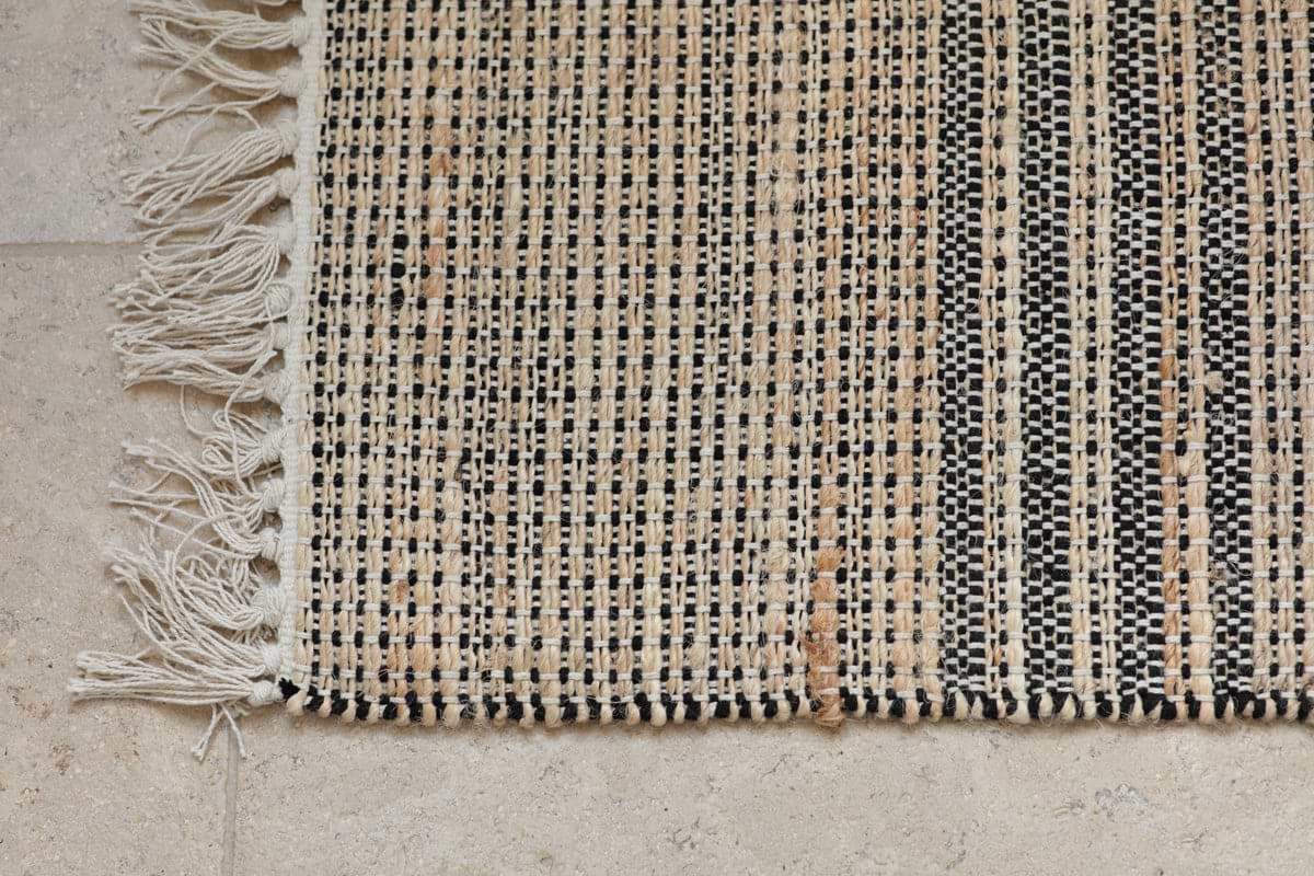 A close-up of the nkuku Varli Jute & Cotton Doormat - Natural shows black, beige, and tan checkered stripes with an off-white fringe on the left edge, resting on a light textured stone floor. The weaving looks coarse and handmade.