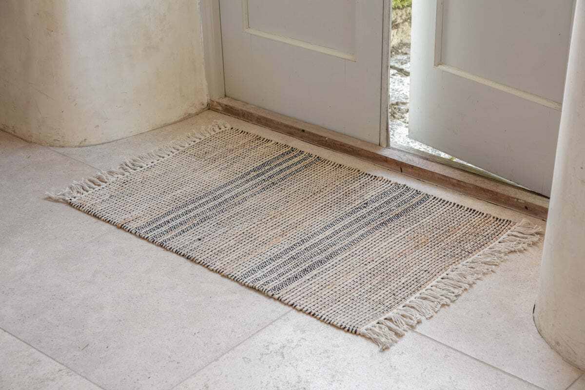 The nkuku Varli Jute & Cotton Doormat - Natural, featuring beige and gray stripes with fringed edges, sits on a light stone floor by a partially open white door as natural light brightens the entryway.