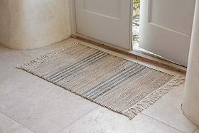 The nkuku Varli Jute & Cotton Doormat - Natural, featuring beige and gray stripes with fringed edges, sits on a light stone floor by a partially open white door as natural light brightens the entryway.