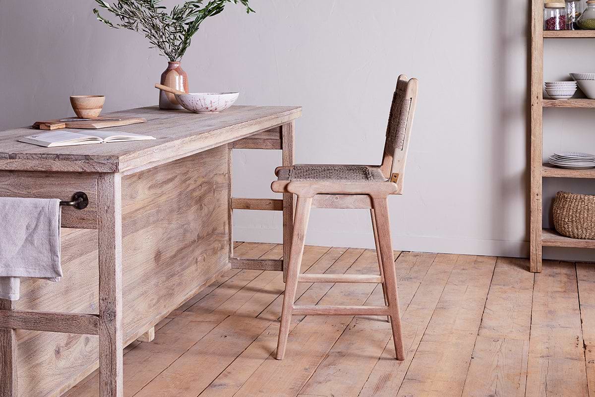 The nkuku Vinay Woven Counter Chair - Natural pairs perfectly with a rustic mango wood kitchen island featuring shelves and a towel hook, offering functional charm and style for your space.