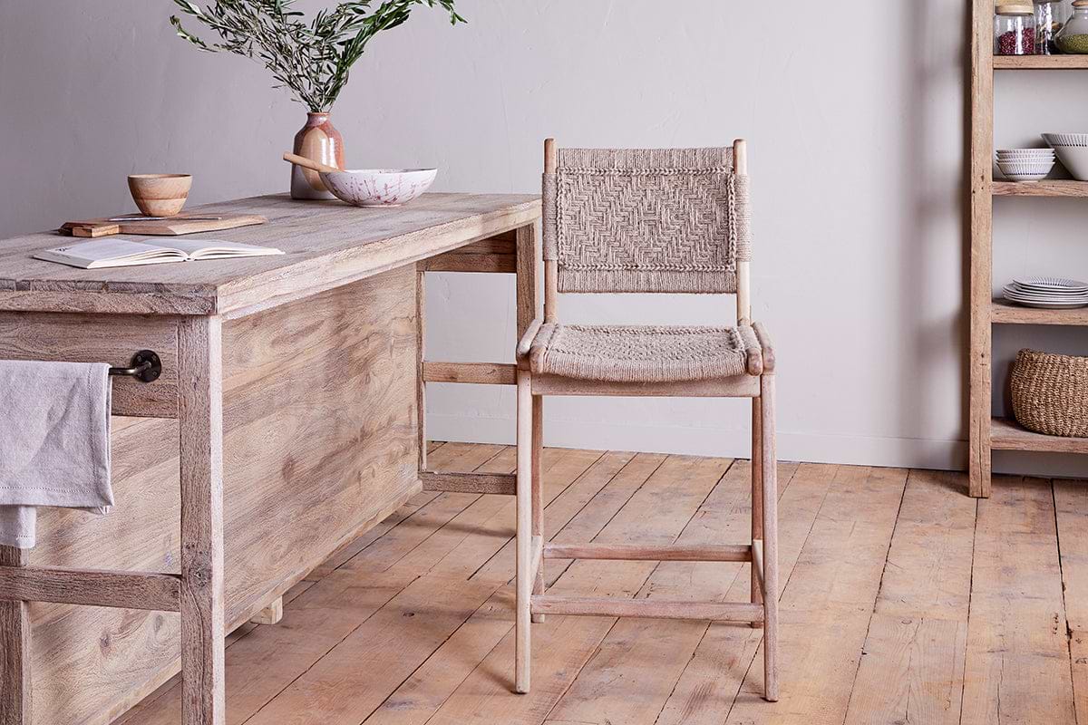 A rustic kitchen features a light mango wood table, nkuku's Vinay Woven Counter Chair - Natural, open shelves with dishes, a wooden floor, and a ceramic vase with greenery. An open book, bowl, and mug add to the cozy minimalist vibe.