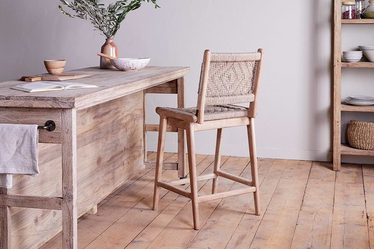 A rustic wooden kitchen showcases the Vinay Woven Counter Chair by nkuku at a mango wood island topped with a vase of green branches, bowl, wooden cup, and notebook. Shelves with dishes and baskets line the wooden floor behind.