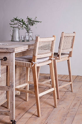 Two nkuku Vinay Woven Counter Chairs in natural mango wood with woven backs and seats are placed at a counter. The counter displays clear glassware and a small vase with green branches, against a white wall and light wood floor.