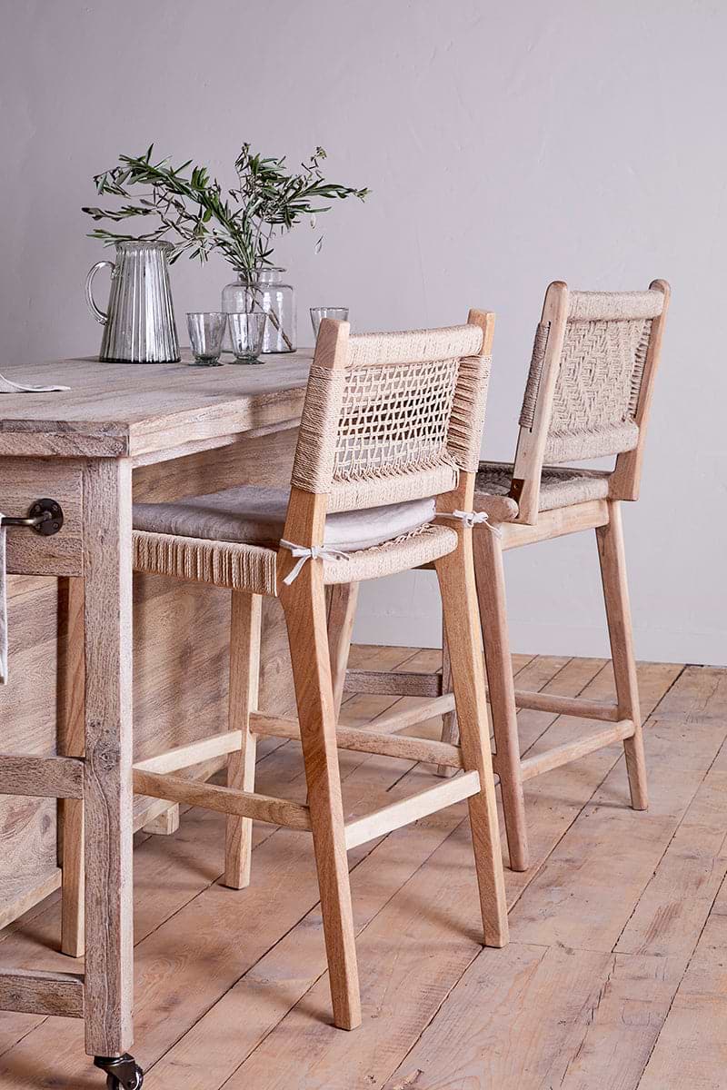 Two nkuku Vinay Woven Counter Chairs in natural mango wood with woven backs and seats are placed at a counter. The counter displays clear glassware and a small vase with green branches, against a white wall and light wood floor.