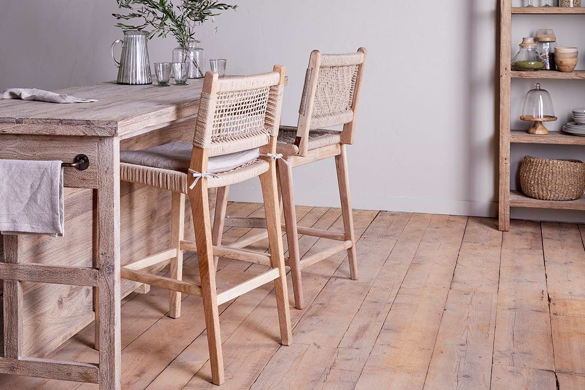 A light mango wood kitchen island is paired with two nkuku Vinay Woven Counter Chairs in natural, set on a pale wood floor. The countertop features a carafe, glasses, and greenery, with a shelf behind displaying dishes, bowls, and baskets.