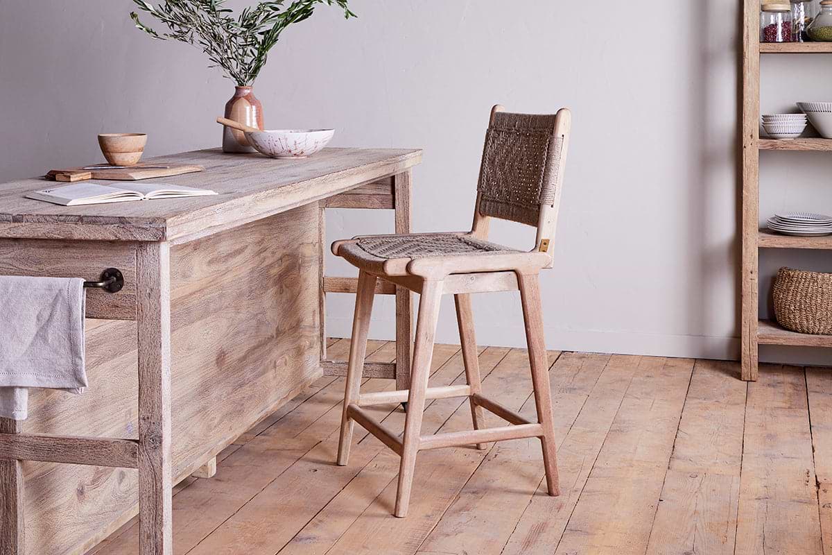 A rustic kitchen with light wood floors features a mango wood island, a Vinay Woven Counter Chair by nkuku, a ceramic jug with green branches, a bowl, and an open book. Shelves display dishes and baskets against a white wall.