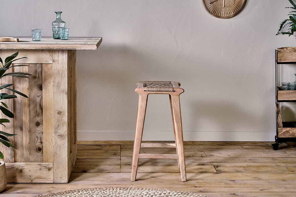 The Vinay Woven Counter Stool - Natural by nkuku, with a jute seat and acacia wood frame, is placed at a rustic wooden bar in a room with plank flooring and a textured wall finish.