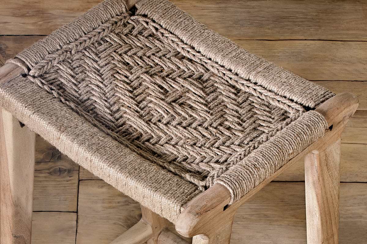 A close-up of the Vinay Woven Counter Stool by nkuku, featuring a natural jute seat in a herringbone pattern and four sturdy acacia wood legs, set on a weathered wooden floor.
