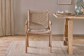The nkuku Vinay Woven Dining Chair - Natural sits on a textured beige rug beside a wooden dining table set with dishes and glassware, surrounded by neutral decor, light wood floors, and jute paneling in the background.