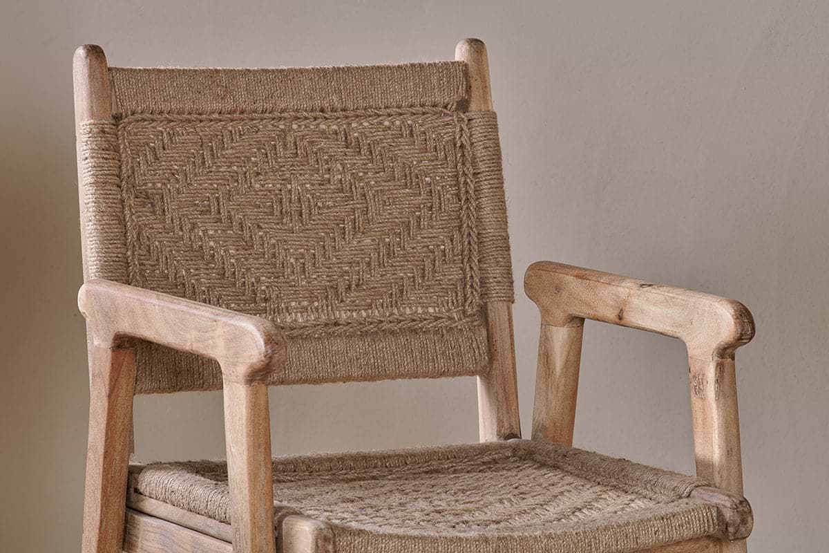 A close-up of the nkuku Vinay Woven Dining Chair - Natural shows its armrests and geometric diamond-patterned jute paneling on the seat and backrest. Crafted from light, unfinished Acacia Wood, it sits against a plain, neutral background.