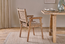 The Vinay Woven Dining Chair - Natural from nkuku, featuring an acacia wood frame and woven seat, pairs with a light wood dining table topped with a glass vase, white napkin, and bowl. Beige curtains and jute paneling add warmth to the cozy room.