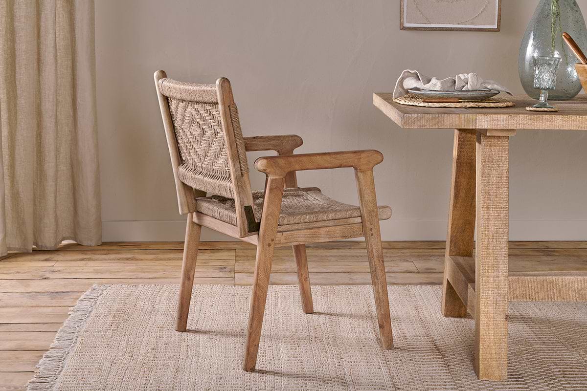 The Vinay Woven Dining Chair - Natural from nkuku, featuring an acacia wood frame and woven seat, pairs with a light wood dining table topped with a glass vase, white napkin, and bowl. Beige curtains and jute paneling add warmth to the cozy room.
