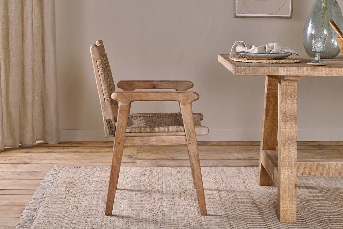 The Vinay Woven Dining Chair by nkuku, featuring a natural acacia wood frame and woven seat and backrest, stands beside a matching table on a beige rug with jute paneling, light beige curtains, and framed artwork in the background.
