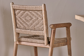 The Vinay Woven Dining Chair - Natural by nkuku features a geometric diamond-patterned jute seat and backrest, set in a light-tone Acacia Wood frame with a smooth finish, shown beside a beige wall and wooden table edge.