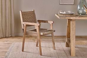 The Vinay Woven Dining Chair by nkuku, featuring a jute seat and backrest, sits next to a textured wooden dining table in a cozy room with light wood floors, a beige rug, and minimal tableware, adding warmth and earthy charm.