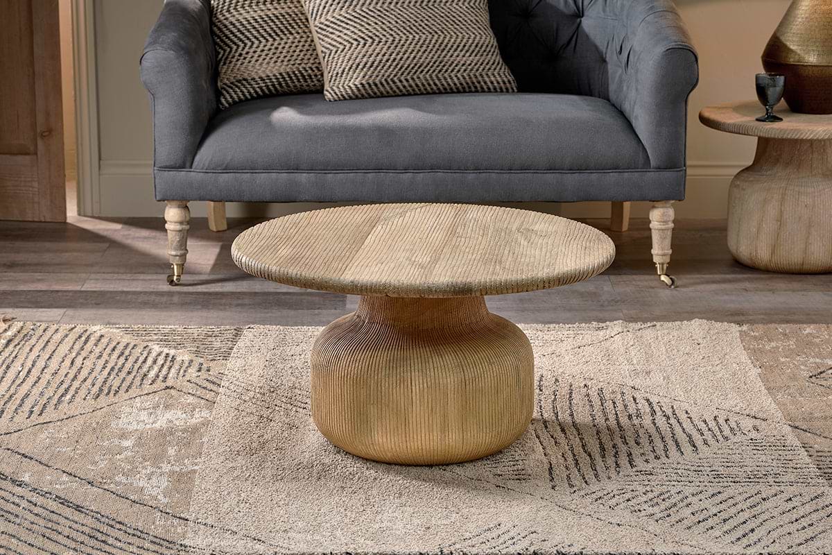 A gold, round nkuku Vivan Grooved Wood Coffee Table with a textured base and smooth top sits on a patterned beige rug, paired with a gray upholstered sofa and patterned pillows for a cozy, modern living space in neutral tones.