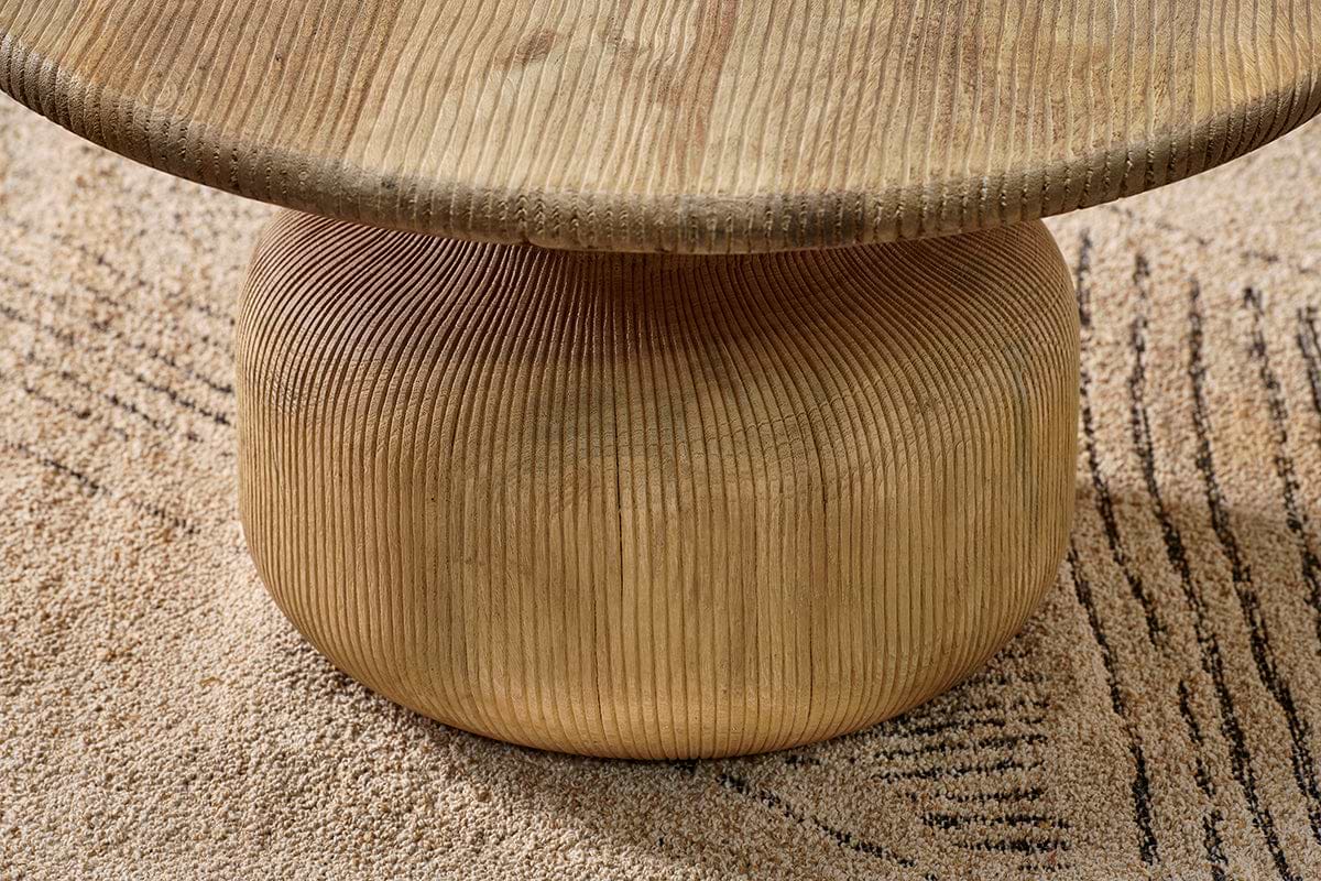 A close-up of the Vivan Grooved Wood Coffee Table by nkuku shows its vertically grooved, rounded base in sustainable mango wood supporting a thick top. The modern table stands on a beige rug with an abstract linear design.