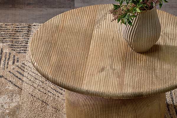 The nkuku Vivan Grooved Wood Coffee Table, crafted from sustainable mango wood, stands on a patterned beige and brown rug, topped with a ribbed ceramic vase and greenery for an earthy touch.