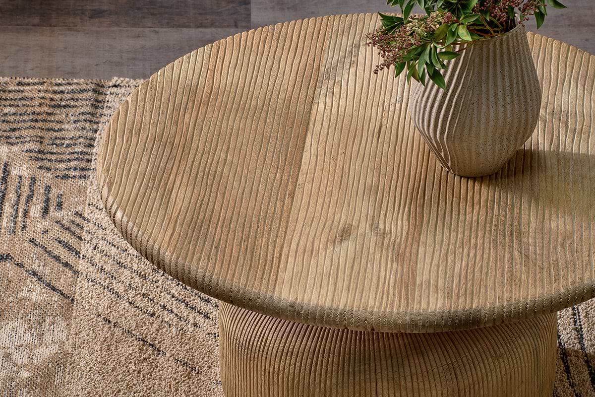The nkuku Vivan Grooved Wood Coffee Table, crafted from sustainable mango wood, stands on a patterned beige and brown rug, topped with a ribbed ceramic vase and greenery for an earthy touch.