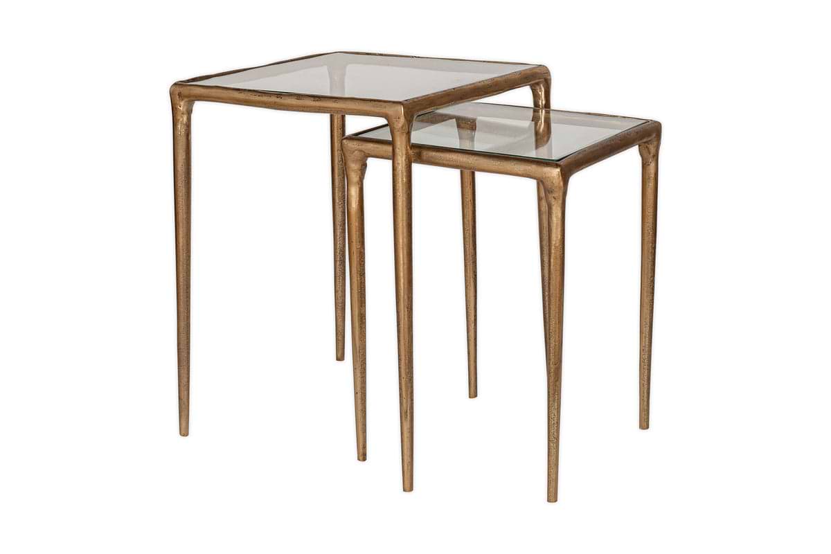 The nkuku Gilasi Cast Metal & Glass Nesting Side Tables (Set of 2) feature slender antique brass frames and clear glass tops. The smaller table slides partially under the larger one, shown against a white background.