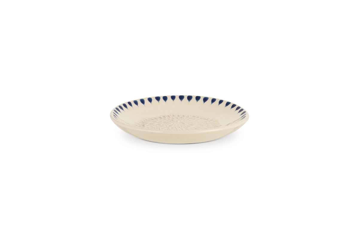 The nkuku Indigo Drop Garlic Grater in Cream & Indigo is a shallow, round beige ceramic plate with a textured center and hand-painted dark blue triangle accents around the rim, perfect for grating garlic.