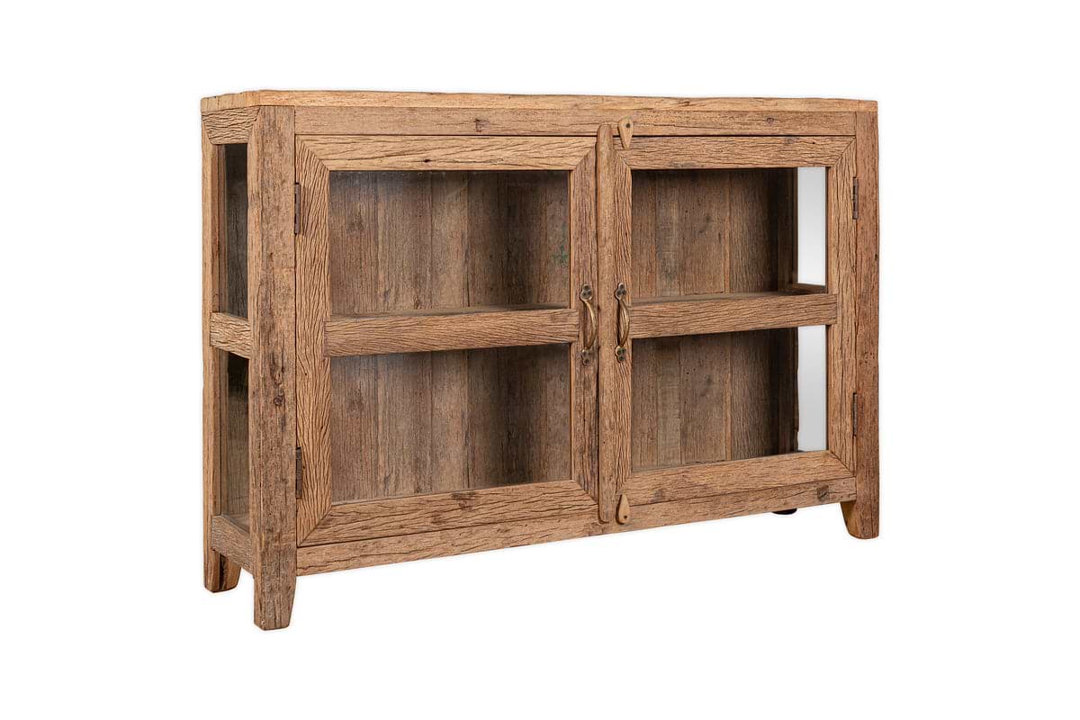 The Ibo Reclaimed Wood & Glass Sideboard by nkuku features two glass-paneled doors with antique brass handles, two interior shelves, a natural weathered finish, and stands on four short legs.