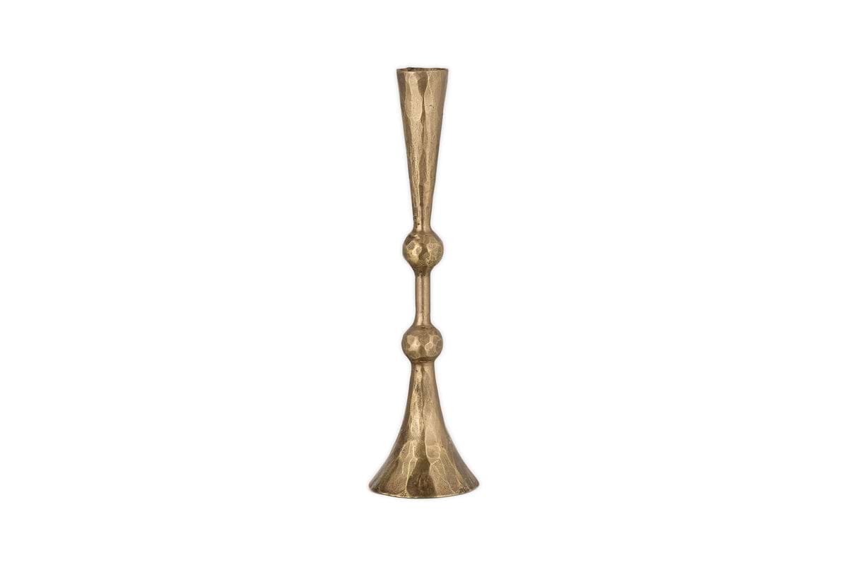 The Javanee Candle Holder by nkuku features a tall, slender silhouette in antique brass with a hammered texture, sculptural elegance, two rounded bulges on its stem, and a flared base. Shown upright against a white background.