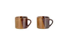 The Naav Mugs - Mustard - Small (Set of 2) by nkuku, each with an angled handle and a glossy mustard and tan color-blocked glaze, are arranged side by side on a white background.