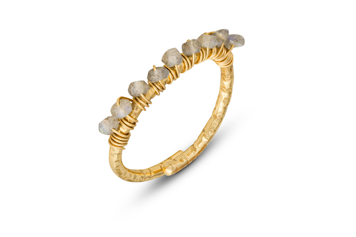 The Vani Labradorite Ring by nkuku features a thin gold-plated band with a textured finish, topped with wire wrap and a row of pale gray labradorite stones, casting a soft shadow on a white background.