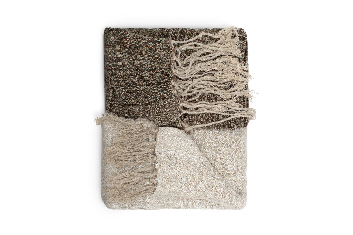 The nkuku Munir Linen Throw - Charcoal features a folded, two-tone design with slubby texture. The top is dark charcoal with matching fringed tassels, while the bottom is lighter beige. Displayed neatly on a white background.