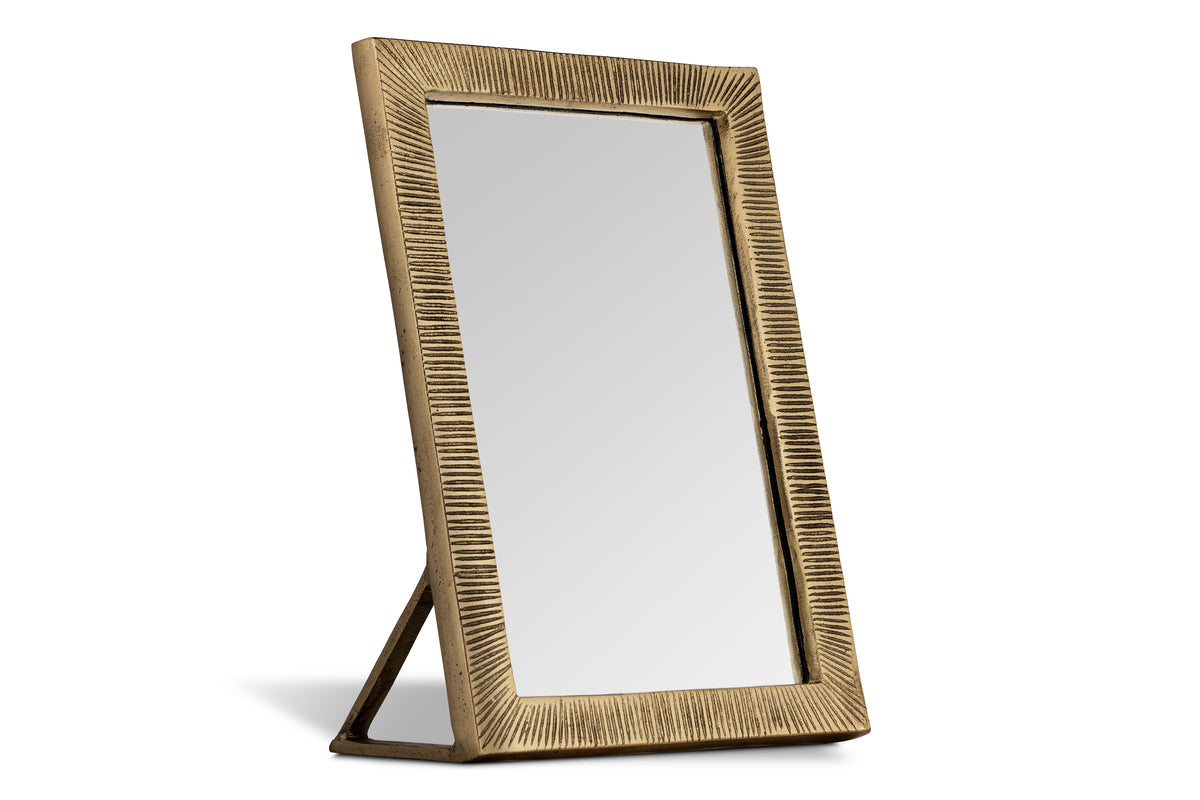 The nkuku Baja Standing Mirror - Antique Brass features a rectangular design with a gold textured frame and vertical grooves. Supported by a triangular stand, it has a modern, elegant look with a metallic matte finish.