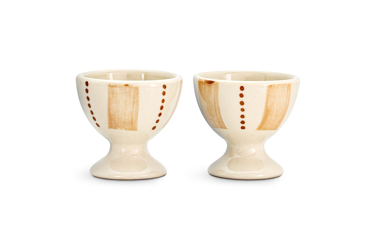 The Patthar Egg Cups - Cream & Clay (Set of 2) by nkuku feature a creamy glaze, vertical tan stripes, and dotted brown lines. Each hand-painted cup has a short, rounded base for added charm. Displayed side by side on a white background.
