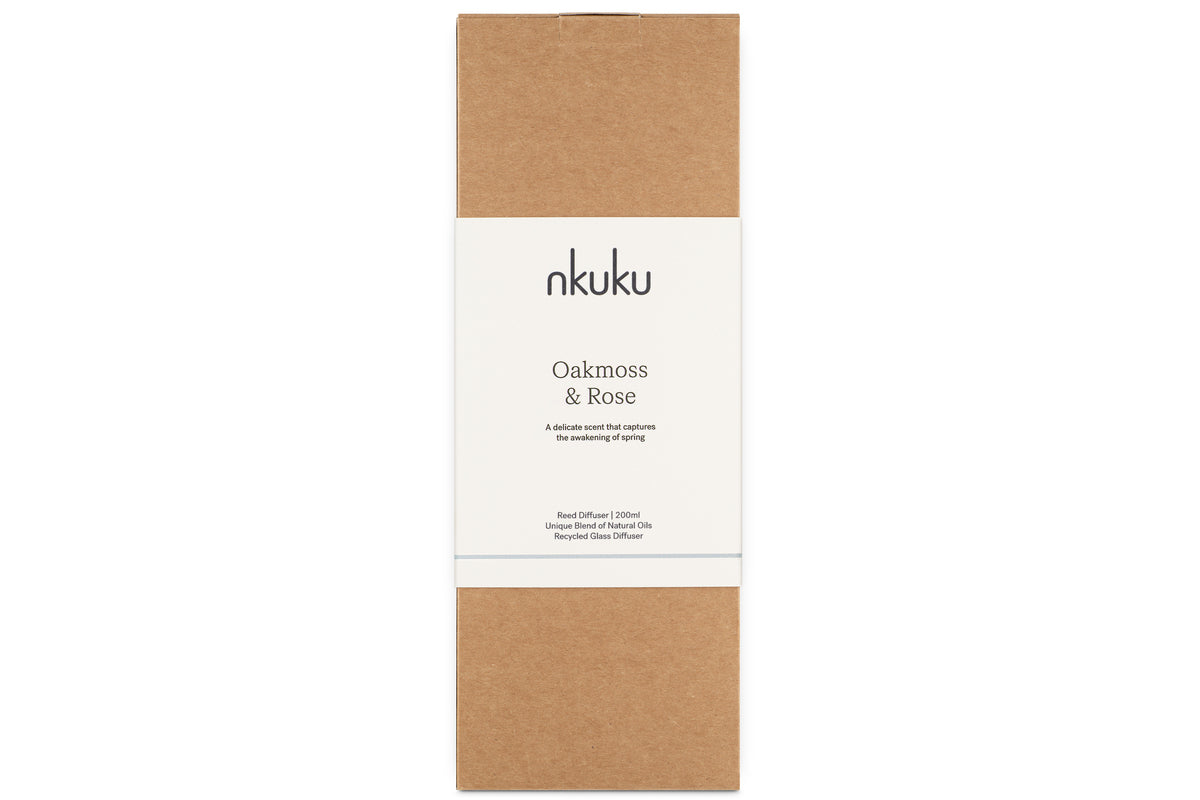 A rectangular brown box with a central white label displaying “nkuku” at the top and “Oakmoss & Rose” below, along with smaller print underneath. The minimal and natural packaging suggests it contains the Oakmoss & Rose Scented Reed Diffuser - Yellow from nkuku, likely featuring a floral fragrance presented in a hand-blown glass vessel.