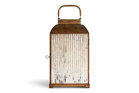 The Ruala Lantern - Brass & Clear by nkuku features a vintage-inspired metal frame in a warm brass finish, clear vertical ribbed glass sides, a top handle for easy carrying, and a side latch for secure closure. Standing upright against a white background, this elegant lantern captures the timeless charm of candlelight.