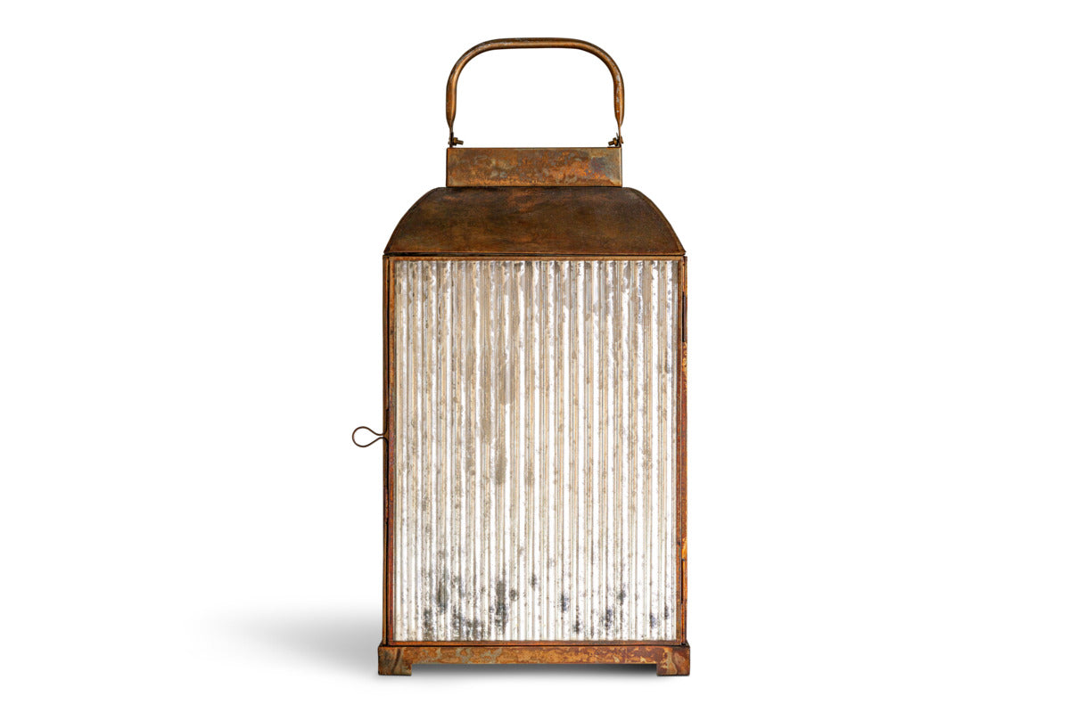 The Ruala Lantern - Brass & Clear by nkuku features a vintage-inspired metal frame in a warm brass finish, clear vertical ribbed glass sides, a top handle for easy carrying, and a side latch for secure closure. Standing upright against a white background, this elegant lantern captures the timeless charm of candlelight.