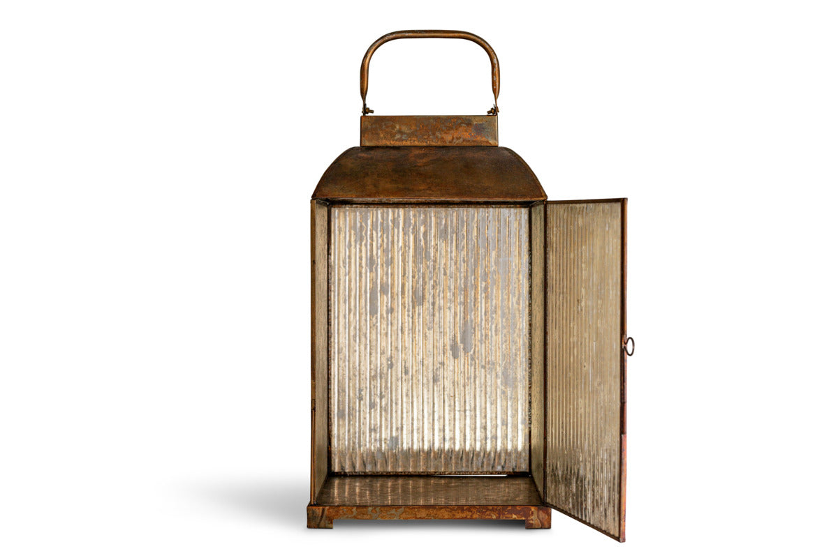 The Ruala Lantern - Brass & Clear by nkuku features a rectangular shape with ribbed clear glass panels and a subtly distressed brass finish. A sturdy metal handle sits on top, while the front door is open to showcase its spacious interior—perfect for adding candlelight. Set against a plain white background.